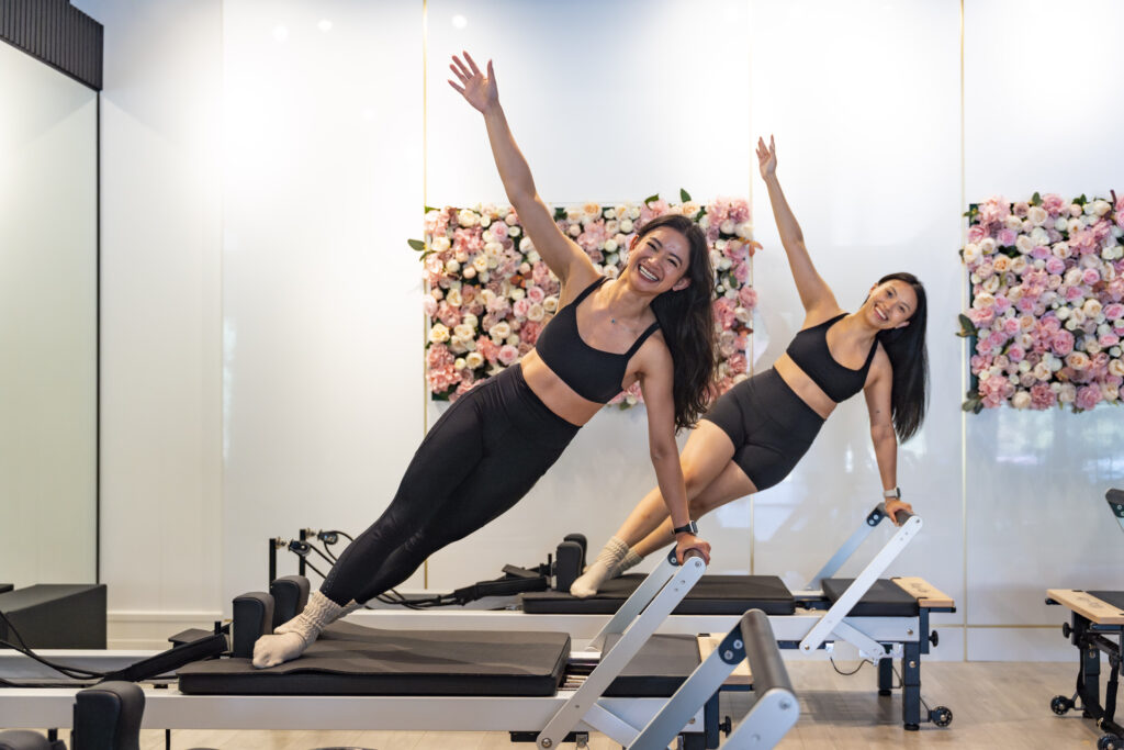 Two women smiling while doing side plank exercises on Pilates reformers in a bright studio with pink floral wall panels.